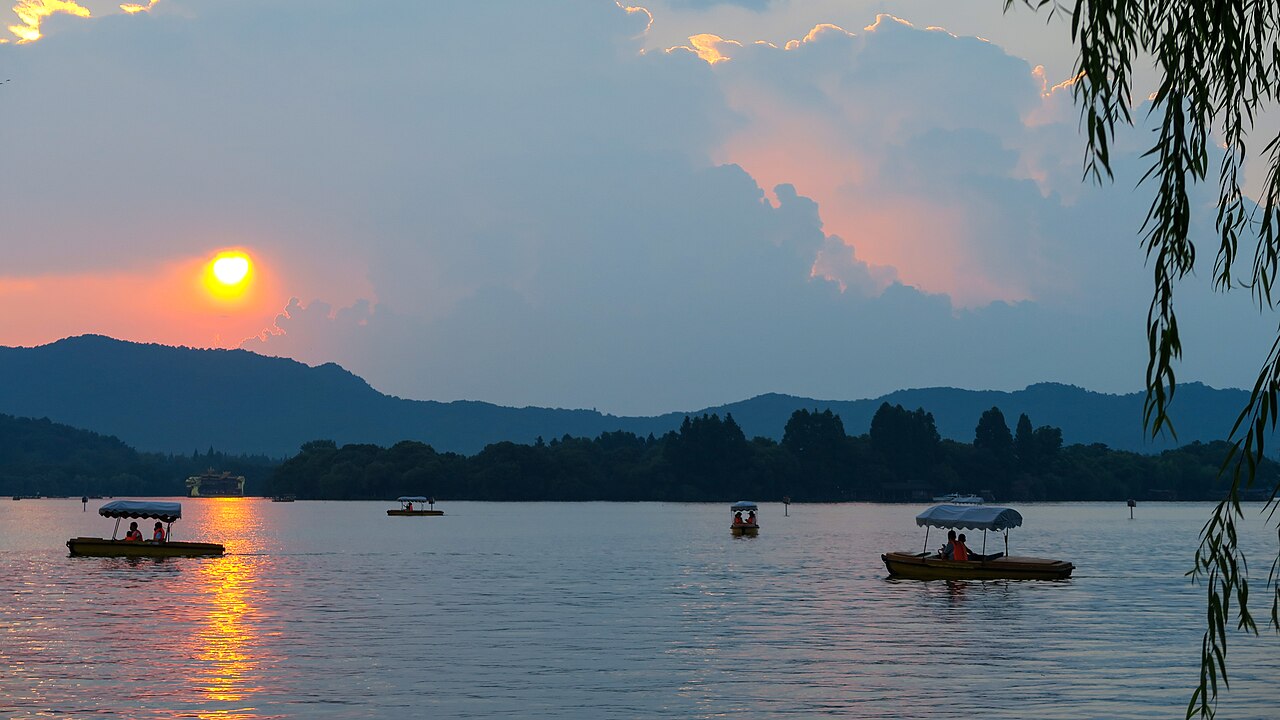 Traditional boats and willow branches on Hangzhou’s West Lake in golden sunset light.